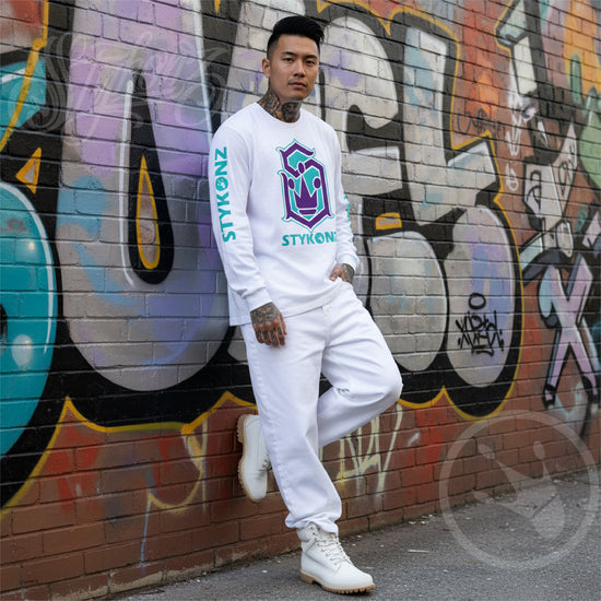 Person wearing a white outfit with 'STYKONZ' branding against a graffiti-covered wall.